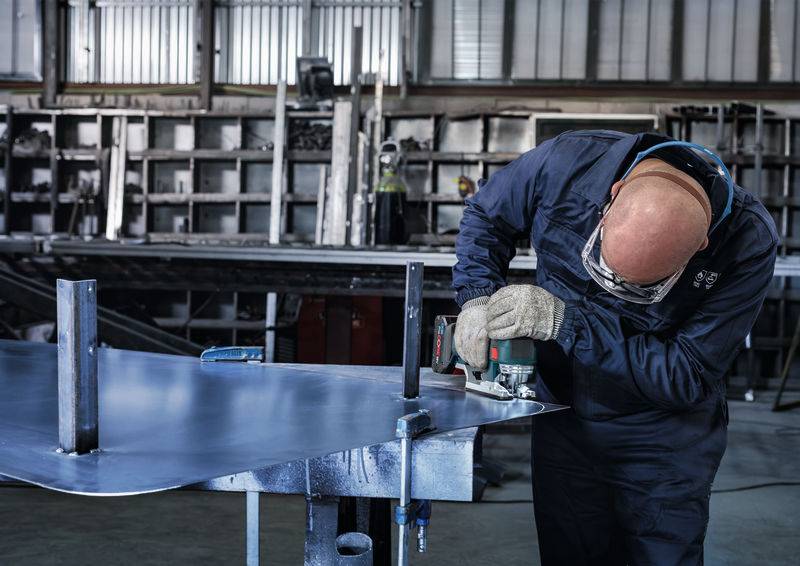 Robotnik w odzieży ochronnej szlifuje kawałek metalu w warsztacie, otoczony przemysłowymi narzędziami i materiałami.