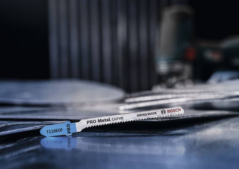 Zbliżenie ostrza do wyrzynarki 'PRO Metal curve' firmy Bosch, leżącego na metalicznym tle, z nieostrymi narzędziami w tle.