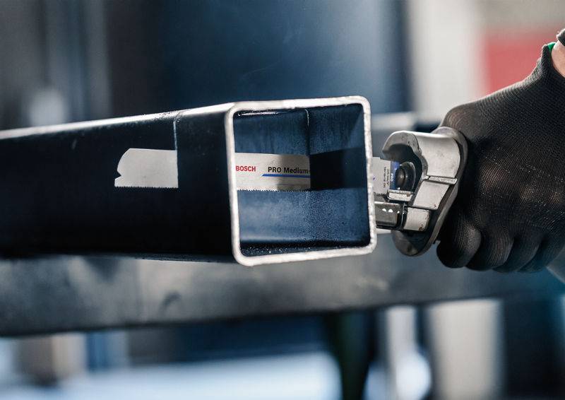 Dłoń w czarnej rękawicy przytrzymuje piłę przy metalowej rurze z naklejką 'Bosch PRO Medium'.