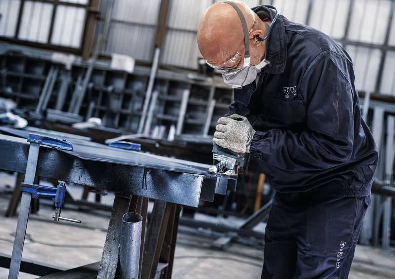 Robotnik w okularach ochronnych i masce używa elektronarzędzia do obróbki metalu w warsztacie. W tle widoczne są regały.