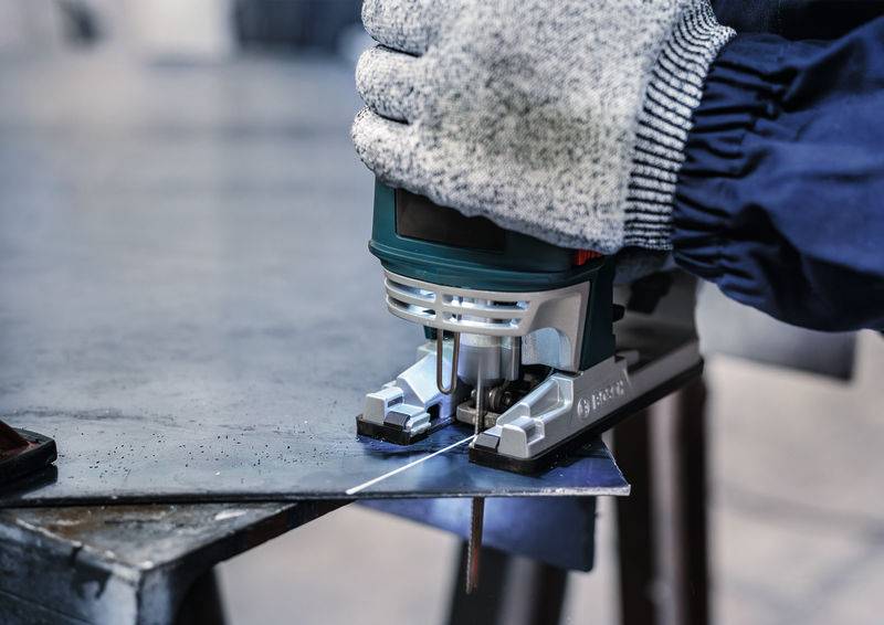 Osoba w rękawicach ochronnych używa wyrzynarki do cięcia płyty metalowej. Fokus na pile i materiale.