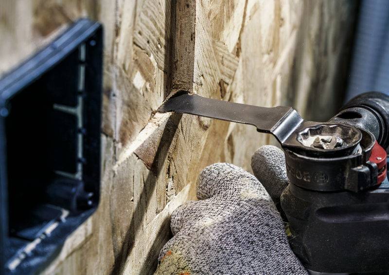 Osoba piłuje elektronarzędziem otwór w desce. Nosi przy tym rękawice ochronne. W tle widać gniazdko elektryczne.