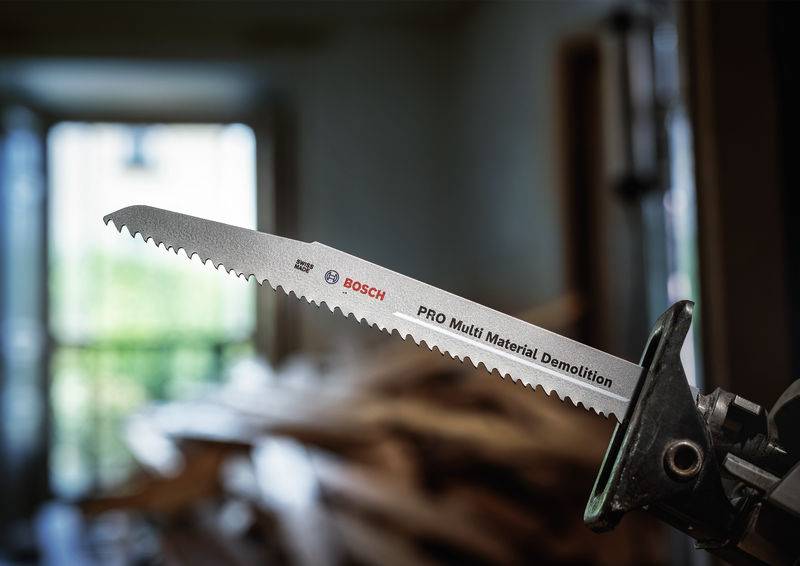 Piła z napisem 'PRO Multi Material Demolition' firmy Bosch w pierwszym planie. Rozmyty tło przedstawia stos drewna.