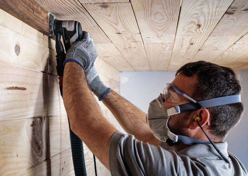 Mężczyzna w odzieży ochronnej szlifuje panele drewniane na suficie za pomocą elektronarzędzia.