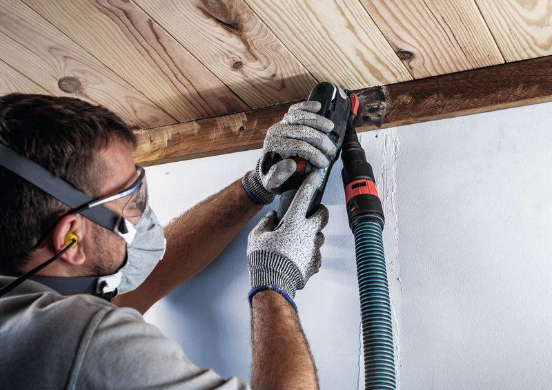 Osoba w okularach ochronnych i masce, szlifująca krawędź drewna elektrycznym narzędziem ściernym przy suficie.