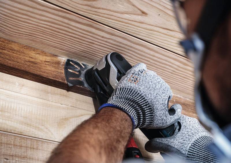 Osoba piłuje drewno elektronarzędziem, nosi szare rękawice ochronne i okulary ochronne. Fokus jest na sytuacji roboczej.