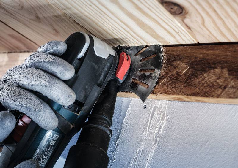 Osoba w szarych rękawiczkach używa elektronarzędzia wielofunkcyjnego do przycinania desek drewnianych przy suficie tak, aby były idealnie równe.