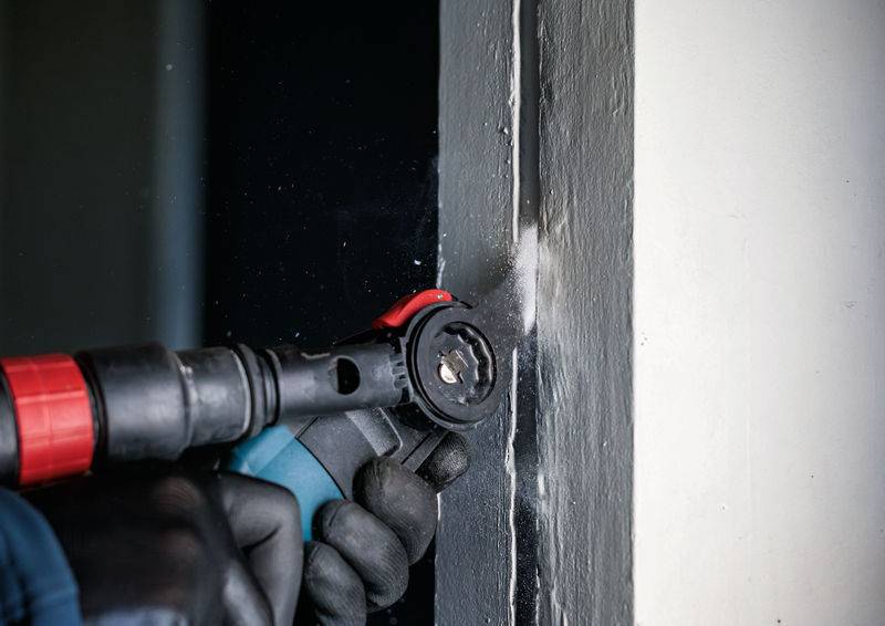 Robotnik w rękawicach używa elektronarzędzia do szlifowania szarej futrymy drzwiowej.