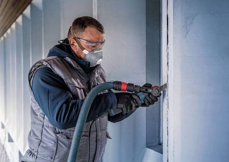 Osoba w odzieży ochronnej używa elektronarzędzia do pracy w betonowej ścianie. Widoczne są okulary ochronne i maska.