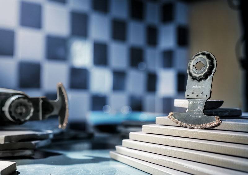 Oscylujące ostrze piły spoczywa na ułożonych kafelkach, w tle widać rozmytą ścianę wyłożoną płytkami.