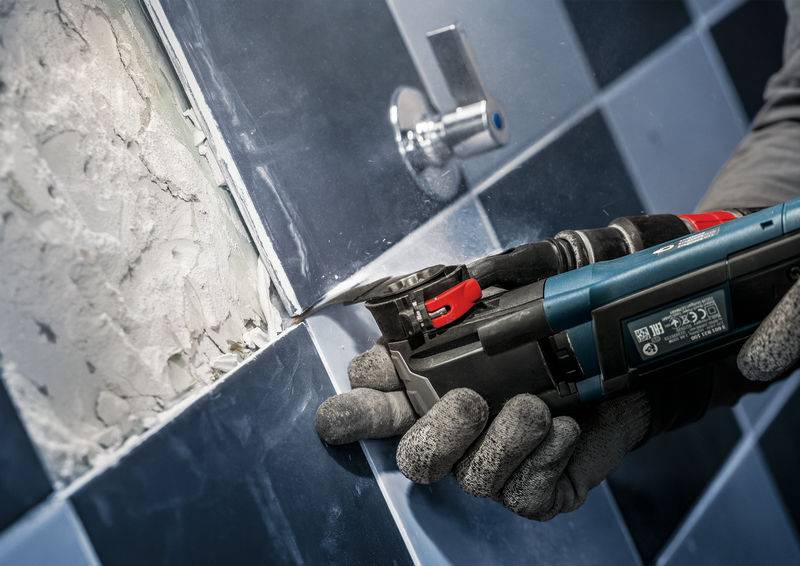 Osoba używa elektronarzędzia z obrotowym ostrzem, aby obrabiać płytki na ścianie.