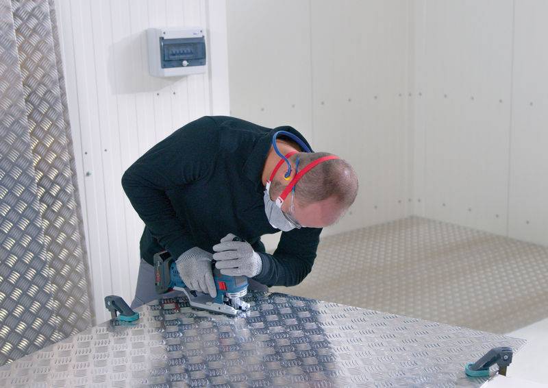 Mężczyzna w masce ochronnej i nausznikach używa piły elektrycznej do cięcia blachy metalowej w warsztacie.