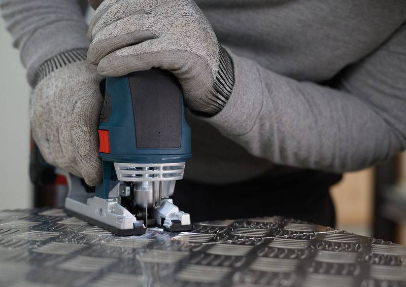 Osoba przecina blachę metalową wyrzynką. Nosi rękawice ochronne.