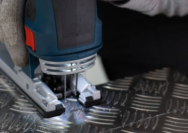 Wyrzynarka przecina arkusz metalu o wzorze rombu. Dłoń w rękawicy pewnie trzyma piłę.