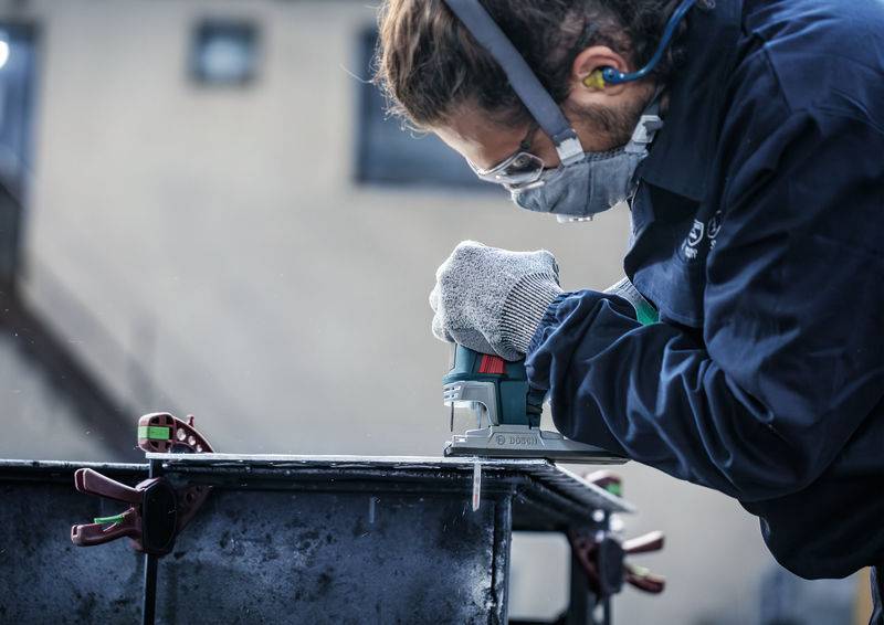 Jedna osoba nosi odzież ochronną i używa piły elektrycznej do cięcia metalu. Otoczenie wskazuje na warsztat.