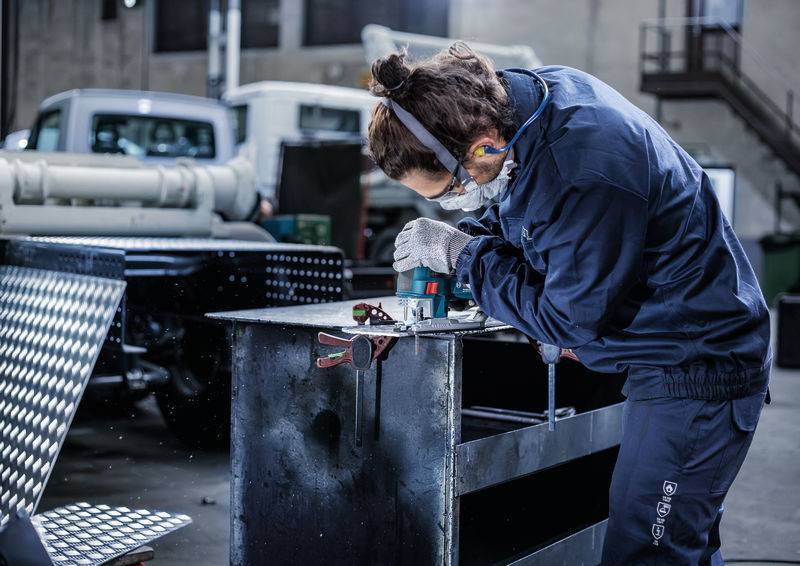 Osoba pracuje w warsztacie przy kawałku metalu, nosząc ochronniki słuchu i okulary ochronne, w otoczeniu pojazdów.