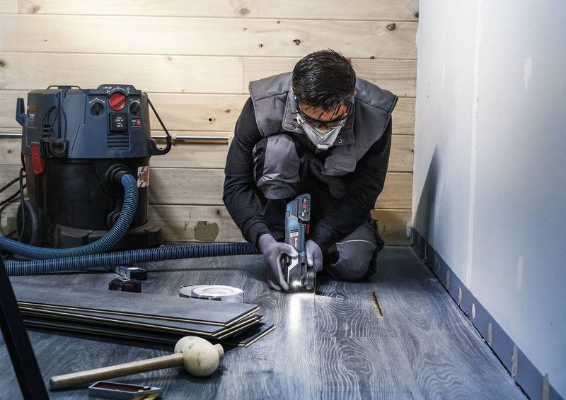 Osoba w okularach ochronnych i masce, klęcząc, pracuje elektronarzędziem na drewnianej podłodze, w tle widoczny odkurzacz.