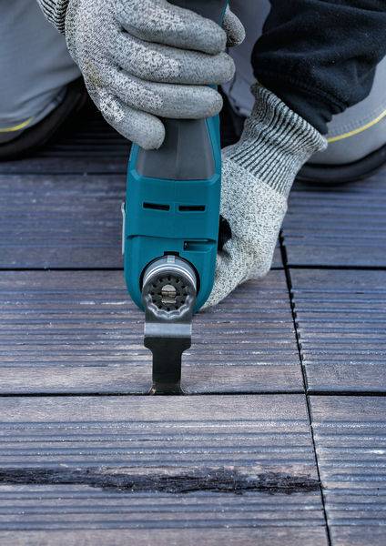 Osoba używa elektronarzędzia do cięcia płyt drewnianych. Zbliżenie rąk w rękawicach roboczych oraz narzędzia leżącego na podłodze.