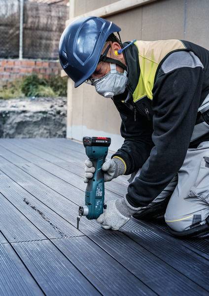 Robotnik budowlany klęczy na tarasie i pracuje elektronarzędziem, ubrany w kask ochronny, maskę i rękawice.