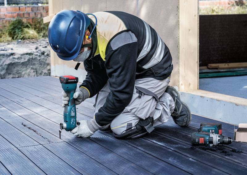 Robotnik budowlany w odzieży ochronnej i kasku mocuje deski drewniane na tarasie za pomocą wkrętarki akumulatorowej na terenie budowy.