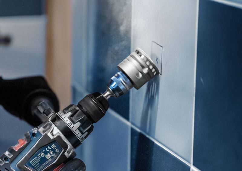 Osoba wierca dziurę w płytce ściennej za pomocą wiertarki. Wiercenie odbywa się w oznaczonym miejscu przy użyciu specjalnej końcówki wiertniczej.