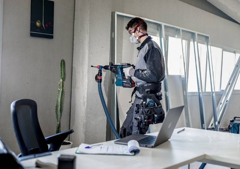 Osoba pracuje elektronarzędziem przy ścianie w biurze, ubrana w odzież ochronną i maskę, na pierwszym planie widoczny jest biurko z laptopem.