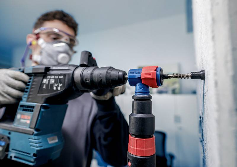 Osoba wierca dziurę w ścianie za pomocą elektrycznej wiertarki. Nosi okulary ochronne, rękawice i maskę ochronną na twarz.