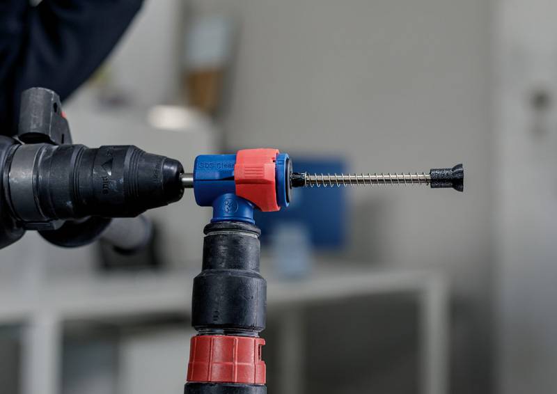 Urządzenie wiertnicze trzyma długą śrubę z czerwono-niebieskim uchwytem. Scena rozgrywa się w środowisku warsztatu.