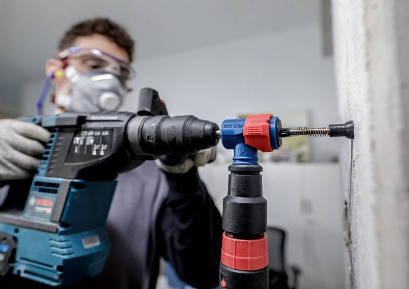 Osoba nosi okulary ochronne i maskę oddechową, używa wiertarki do wiercenia w ścianie betonowej. Bezpieczeństwo jest na pierwszym miejscu.