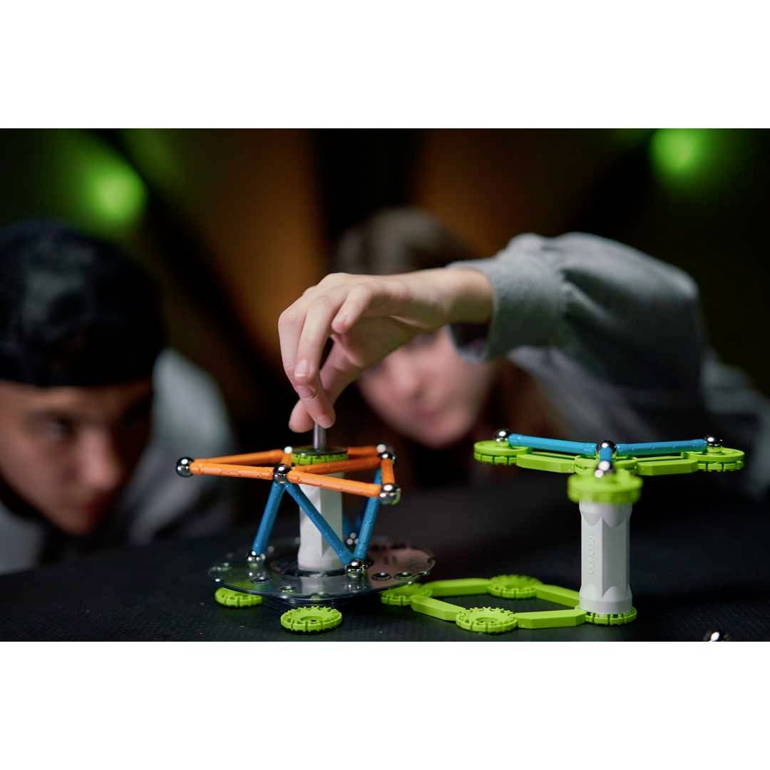 Dwie osoby bawią się kolorowymi klockami. Jedna osoba sięga po pomarańczowy element strukturalny, podczas gdy druga przygląda się jej.