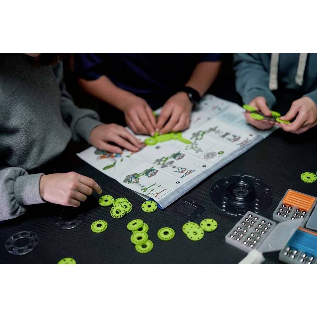 Dwie osoby składają zestaw robotyczny z zielonymi kołkami zębatymi i instrukcją. Na stole leżą różne elementy konstrukcyjne.