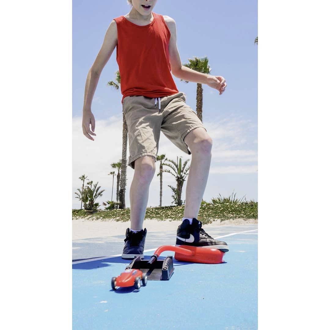 Dziecko w czerwonym tanktopie i beżowych szortach stoi na niebieskim podłożu w pobliżu palm. Deskorolka leży przed nim.