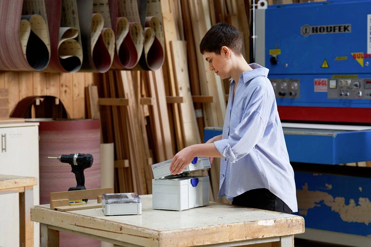 Osoba w warsztacie ubrana w niebieską koszulkę, porządkująca przedmioty wewnątrz pudła. W tle widoczne są wiertarki i drewniane materiały.