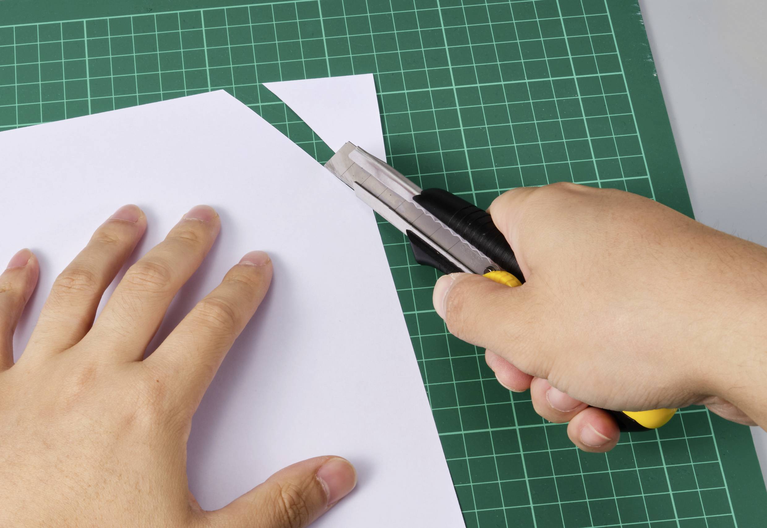 Jedna ręka kroi białą kartkę żółtym nożykiem na zielonej macie do cięcia.