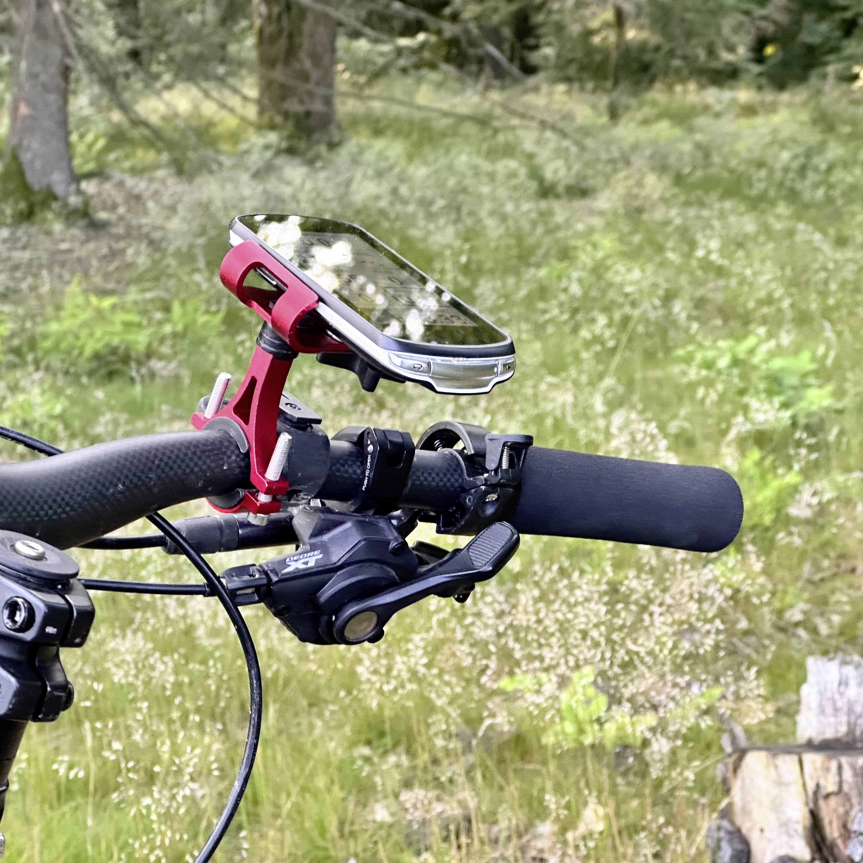 Zbliżenie kierownicy roweru z zamontowanym uchwytem na smartfona w lesie. W tle widoczne są drzewa i trawa.