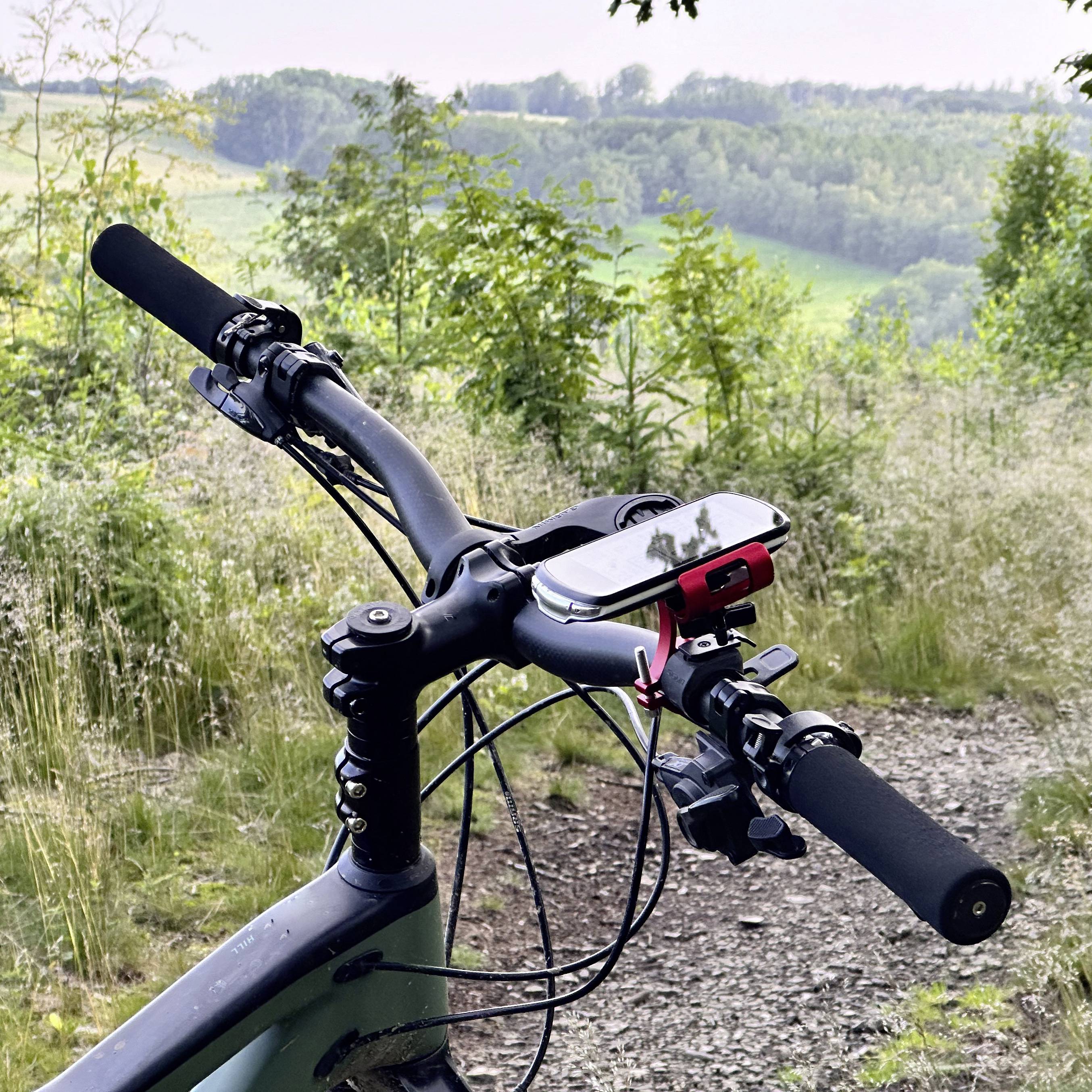 Kierowca roweru z przymocowanym smartfonem, spoglądający na malowniczą leśną drogę, otoczony drzewami i łąkami.