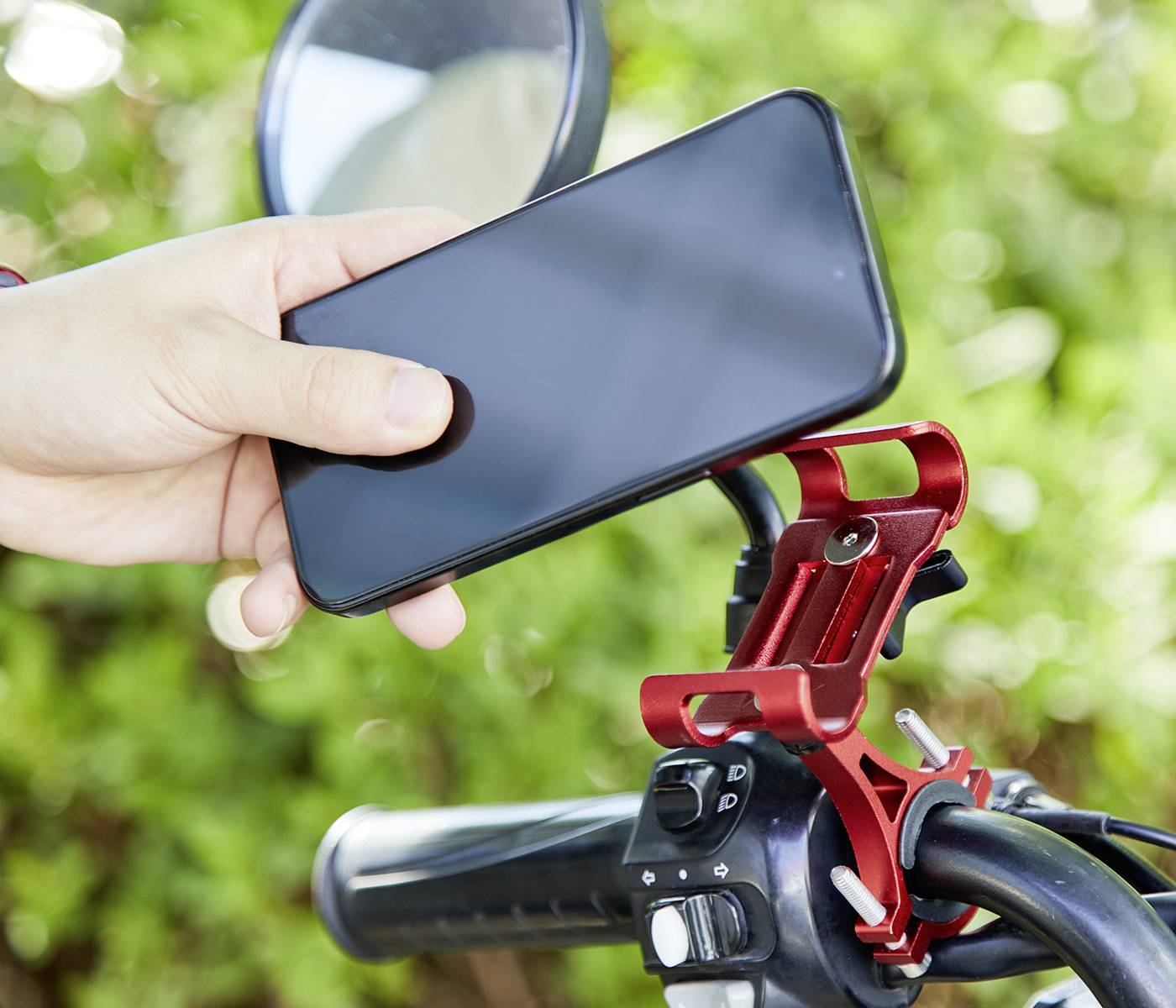 Osoba trzyma smartfon nad czerwonym uchwytem rowerowym zamocowanym na kierownicy roweru. W tle widoczne są zielone liście.