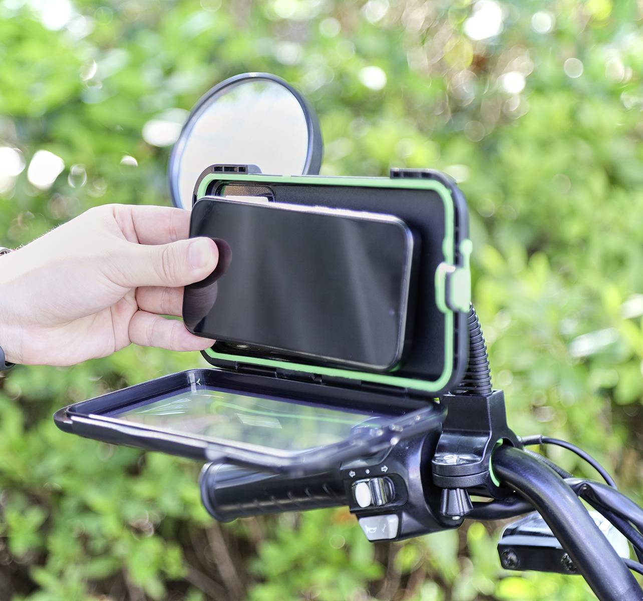 Kierownica roweru z uchwytem na smartfona, do którego człowiek wkłada telefon. W tle nieostre zielone krzewy.