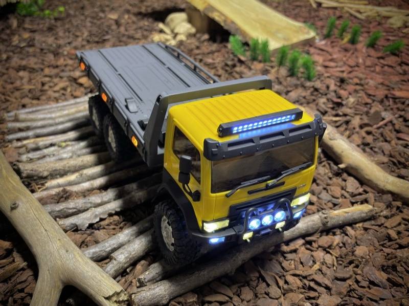 Żółta miniaturowa ciężarówka z jaśniejącymi reflektorami jedzie po nierównej drodze złożonej z kawałków drewna w krajobrazie makiety.