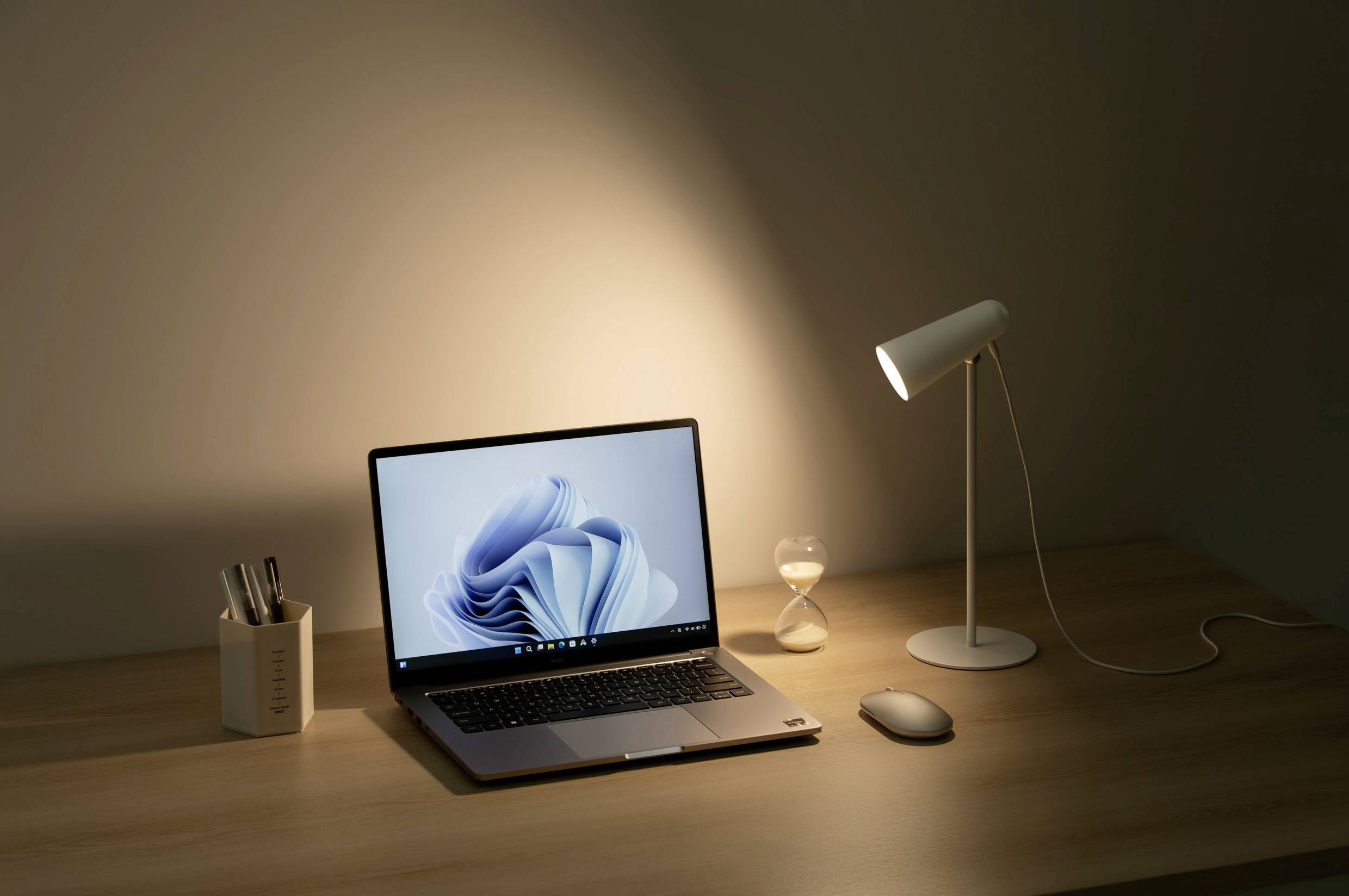 Oświetlone biurko przedstawia otwarty laptop, klepsydrę, lampkę oraz pojemnik na długopisy w delikatnym świetle.