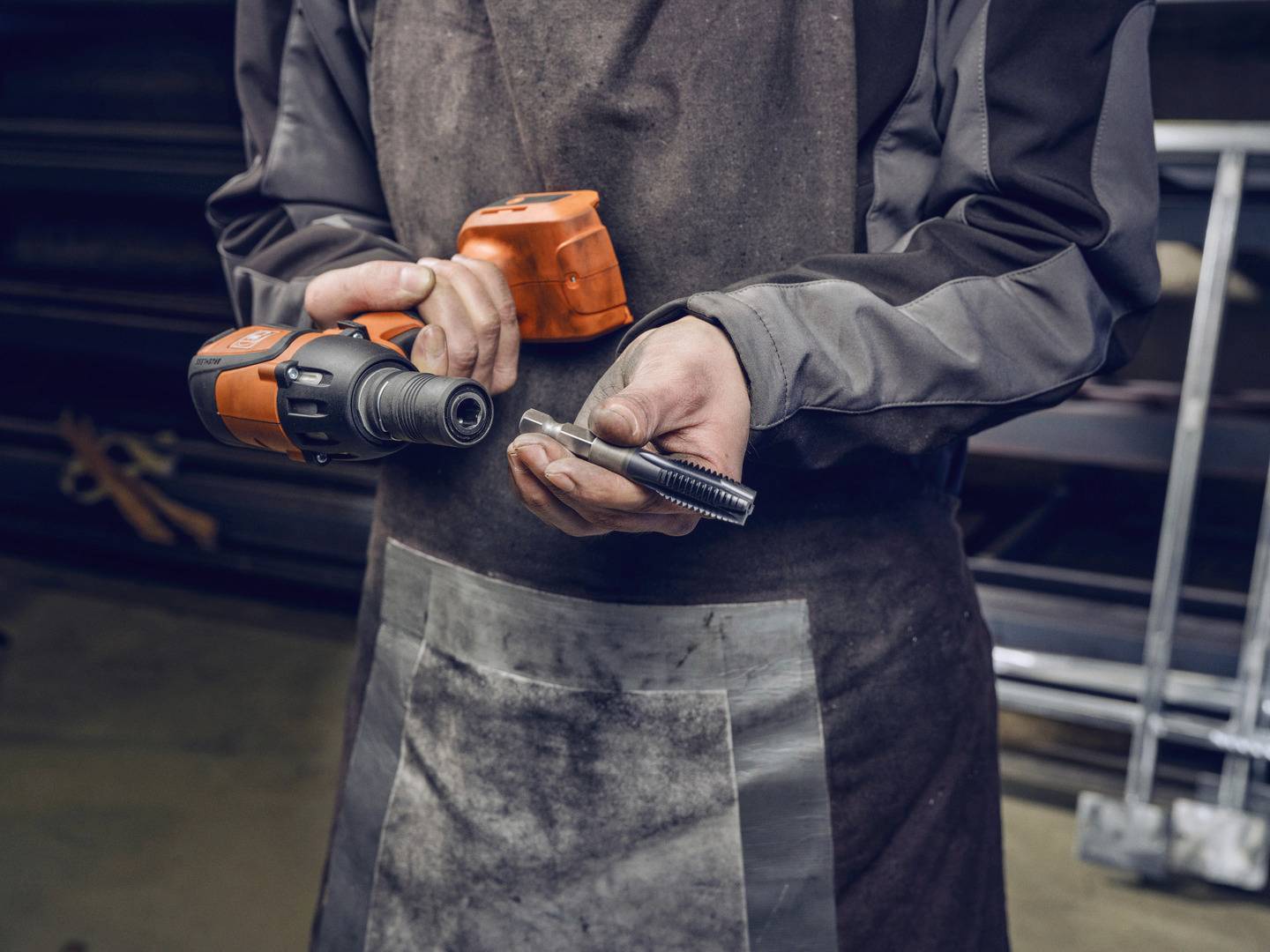 Osoba trzyma w jednej ręce akumulatorowy wkrętak, a w drugiej śrubę, ubrana jest w brązowy fartuch i ubranie robocze.