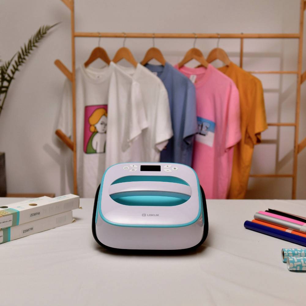 Żelazko stoi na stole przed kolorowymi, zadrukowanymi koszulkami na wieszakach. Obok leżą opakowania i długopisy.