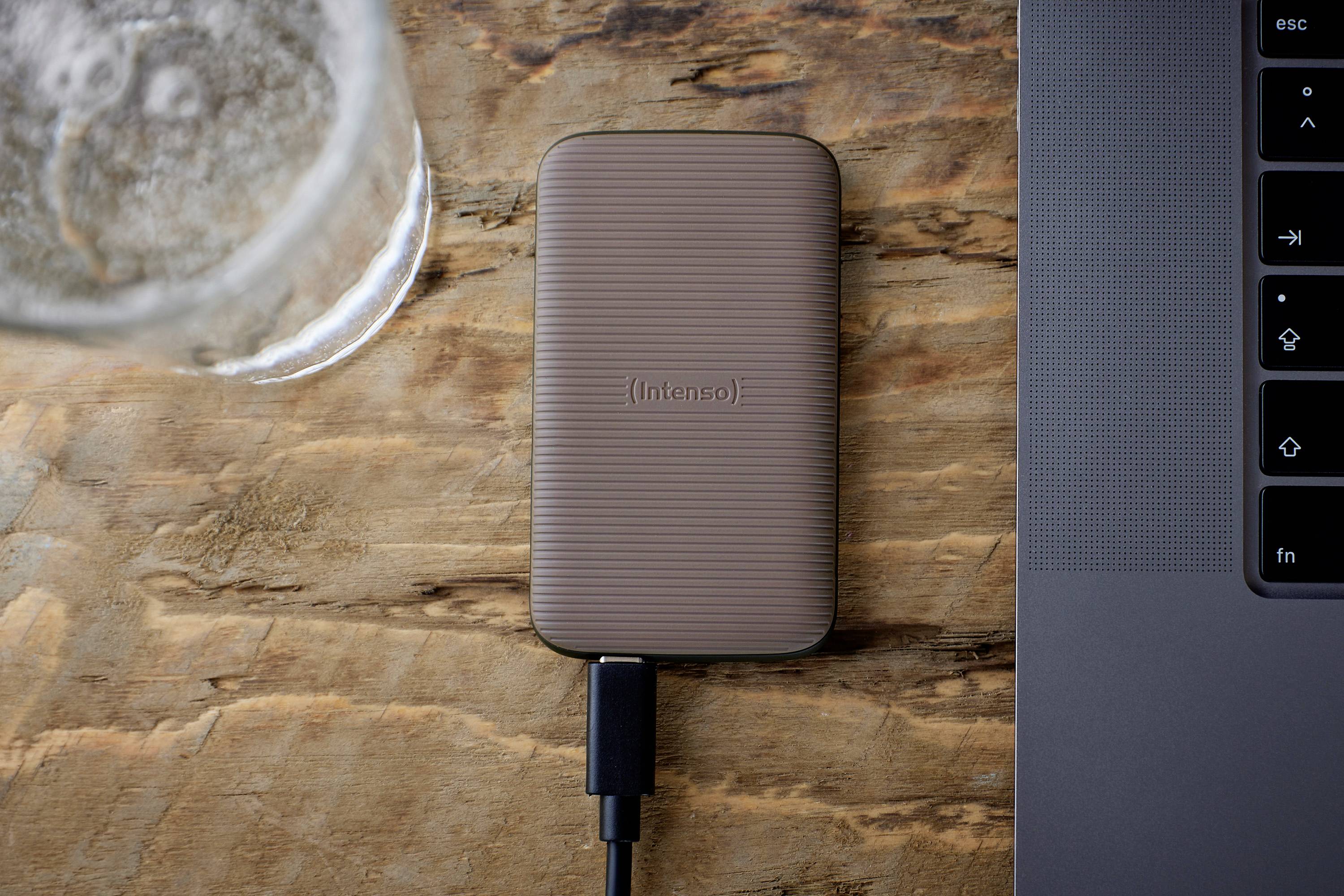 Zewnętrzny dysk twardy jest podłączony do laptopa za pomocą kabla. Na pierwszym planie widać laptopa, a w tle stoi szklanka z napojem na drewnianym stole.