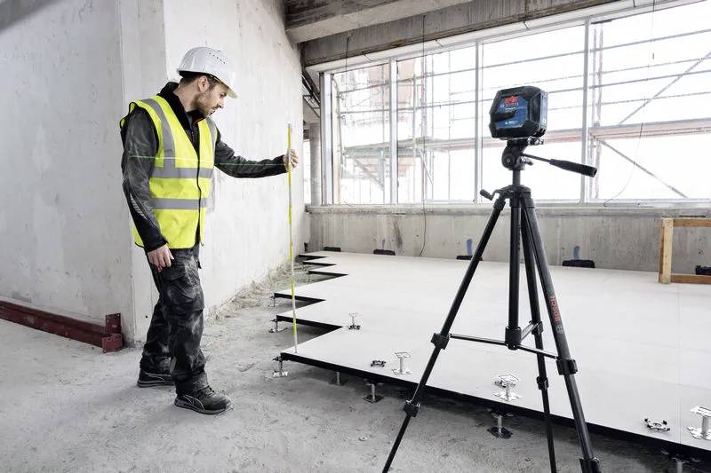 Robotnik budowlany mierzy wysokość podniesionego panelu podłogowego za pomocą miarki. W pobliżu ustawiony jest statyw z poziomicą laserową.