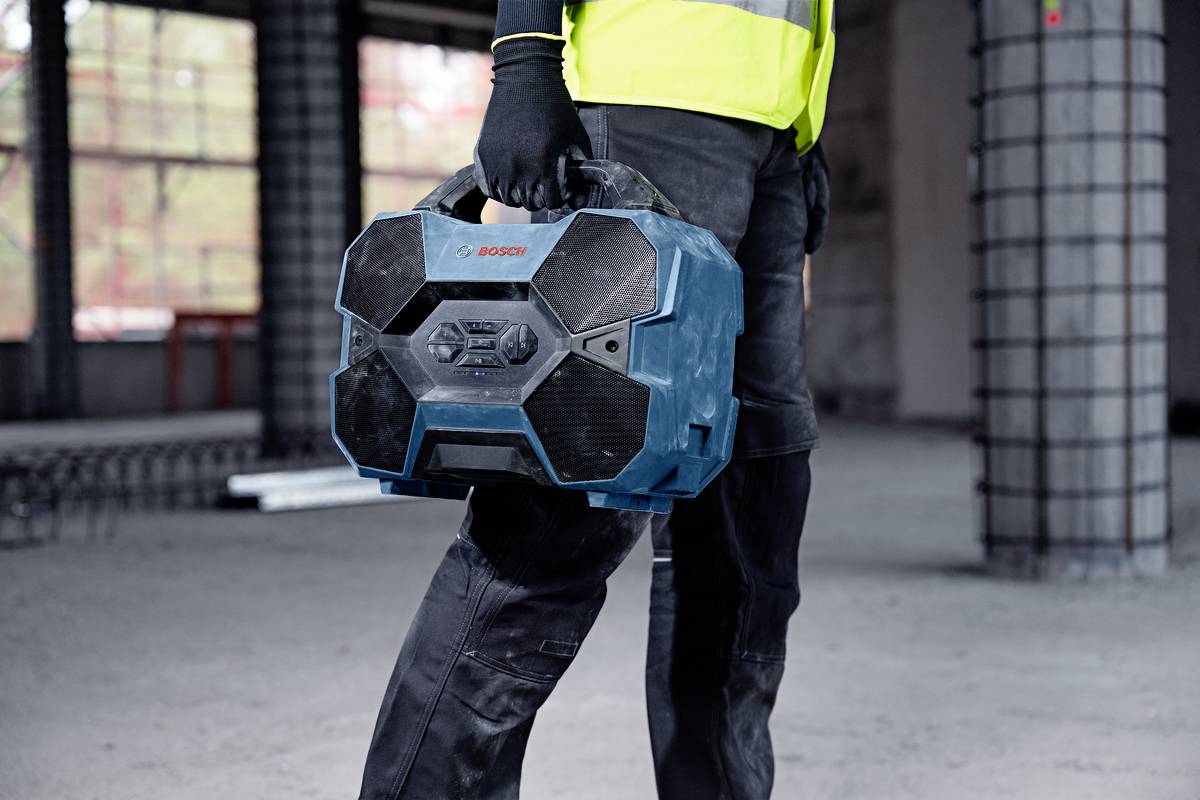 Robotnik budowlany niesie niebieską, sześciokątną skrzynkę narzędziową w niedokończonym budynku. Pracownik ma na sobie kamizelkę odblaskową.