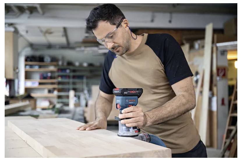 Mężczyzna w okularach ochronnych używa elektrycznej szlifierki na drewnianej powierzchni w warsztacie stolarski, z całkowitym skupieniem wykonując swoją pracę.