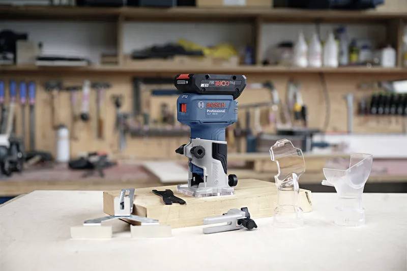 Kompaktowy niebieski i czarny frezar stolarski jest umieszczony na stole warsztatowym, otoczony różnymi przezroczystymi osprzętem i narzędziami. W tle widoczne jest wnętrze warsztatu.