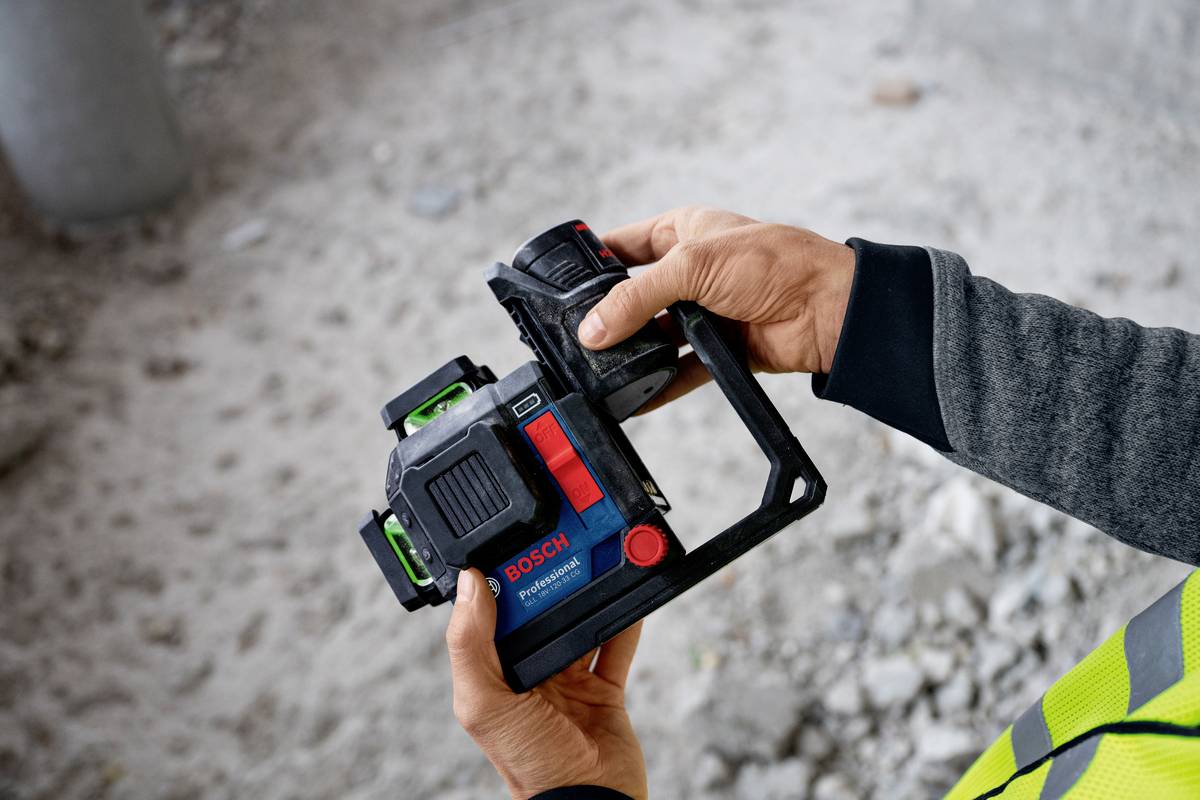 Dwie ręce trzymają kamerę z narzędzi Bosch, używaną do precyzyjnych pomiarów na placu budowy. W tle widoczna jest ziemia i beton.