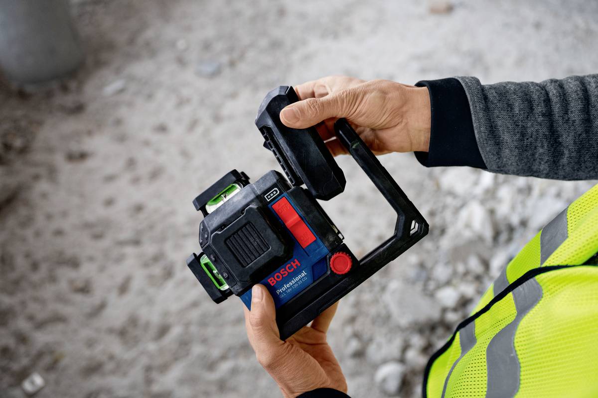 Osoba trzyma urządzenie pomiarowe firmy Bosch z zieloną i czerwoną wskazówką, jest ubrana w kamizelkę odblaskową, w otoczeniu placu budowy.