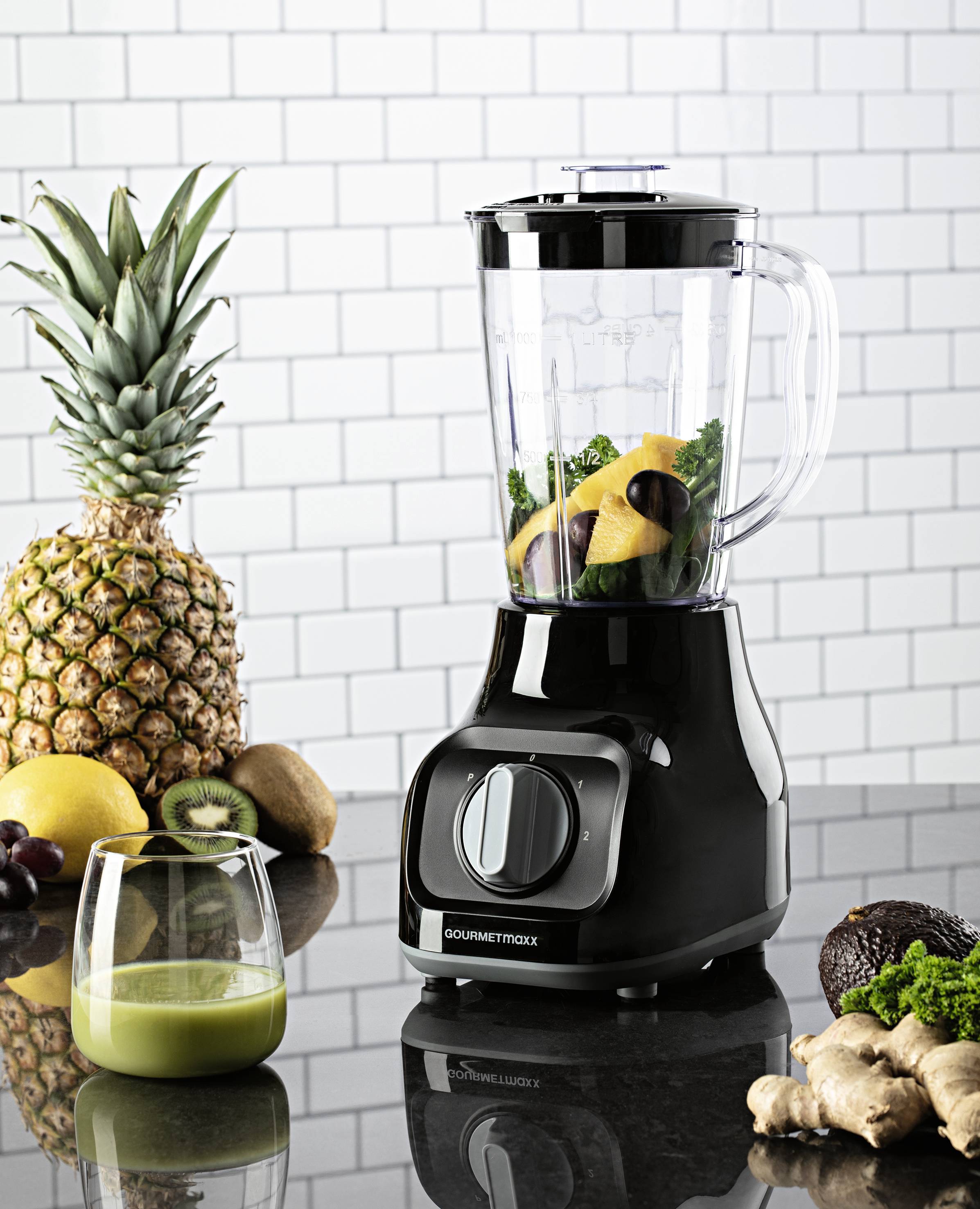 Czarny blender stoi na blacie kuchennym obok owoców takich jak ananas i kiwi. W blenderze znajdują się zielone liście i kawałki owoców.